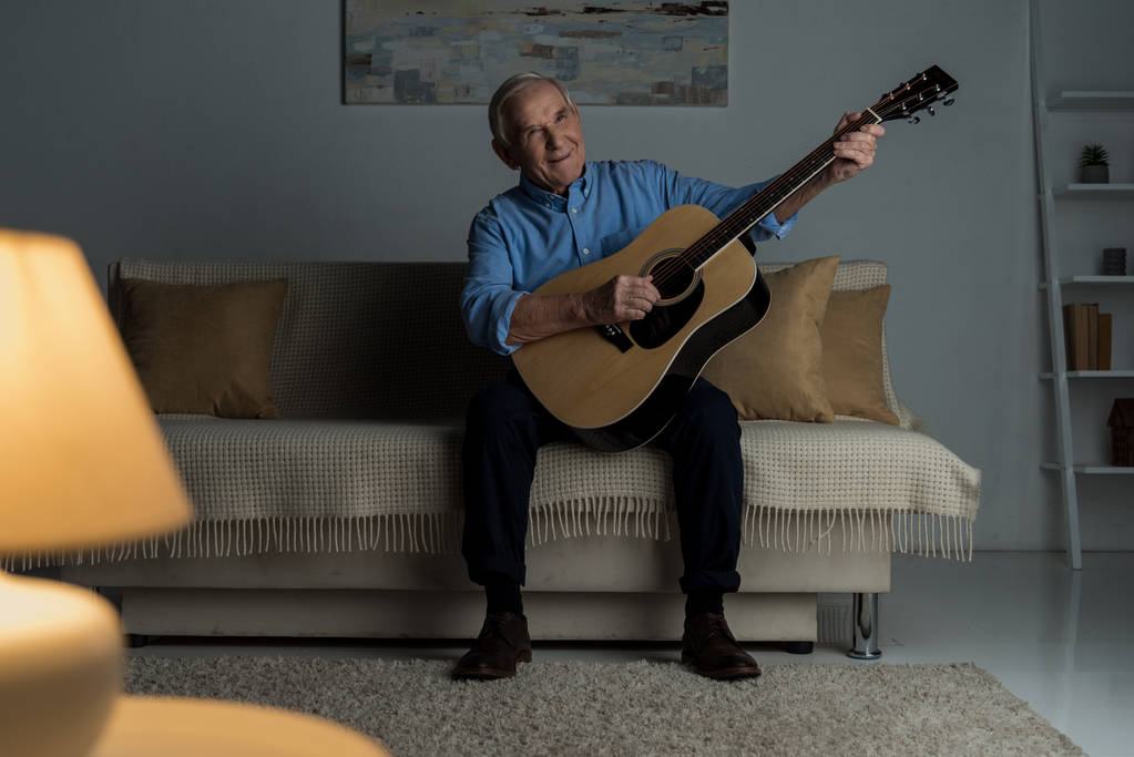 Starszy człowiek szczęśliwy gra gitara akustyczna siedząc na kanapie w pokoju - Zdjęcie, obraz