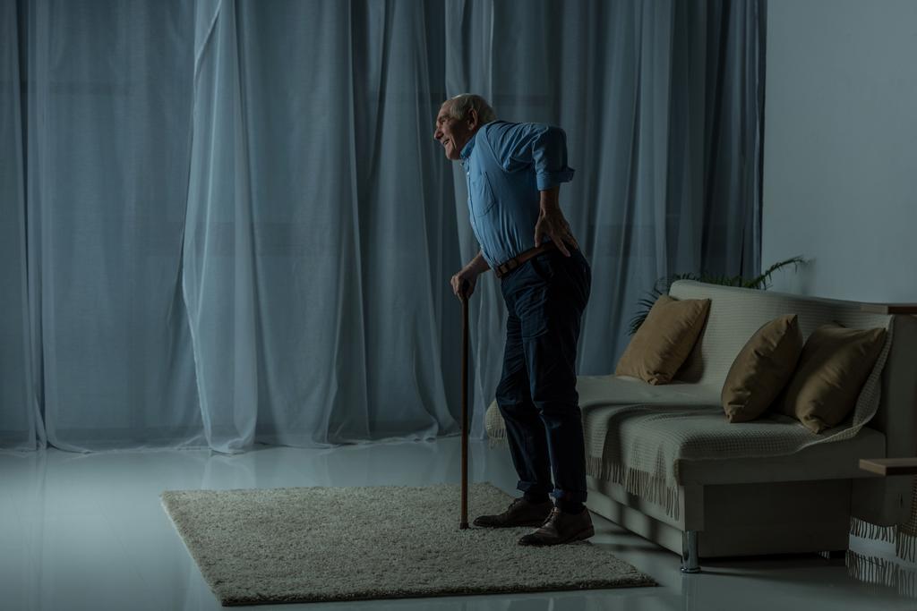 Homem idoso que sofre de dor nas costas se inclina em uma bengala no interior do quarto vazio
 - Foto, Imagem