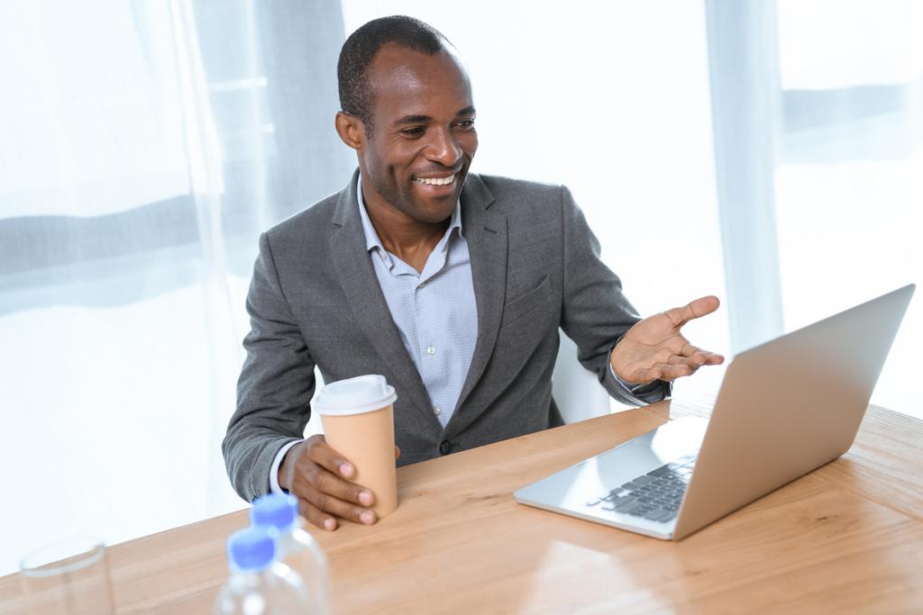 усміхнений африканський чоловік з чашкою кави посміхається, дивлячись на ноутбук на столі
  - Фото, зображення