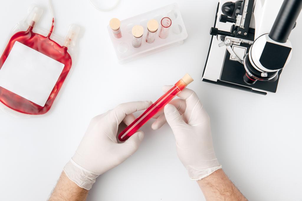 Draufsicht der Hände in Handschuhen mit Blut für Transfusionen in Fläschchen und Mikroskop isoliert auf weißem Hintergrund    - Foto, Bild