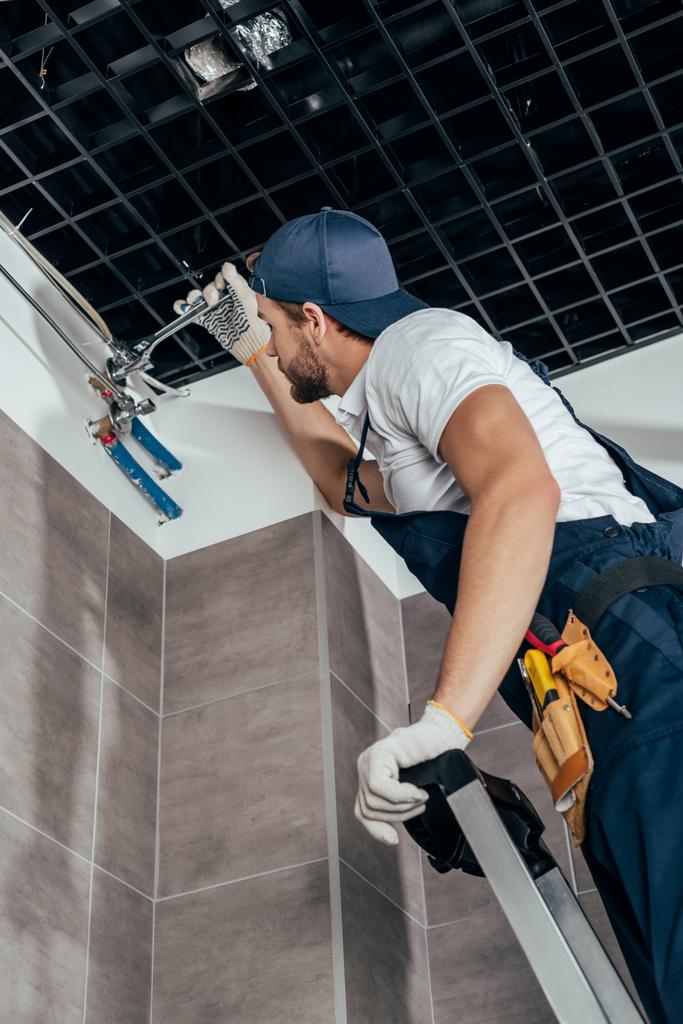 nízký úhel pohledu instalatér, stojící na žebříku a práci s potrubím v koupelně  - Fotografie, Obrázek