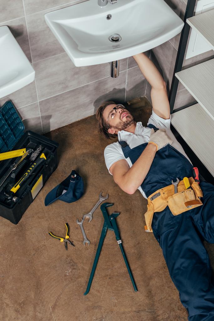 vista ad alto angolo di giovane idraulico maschile che fissa il lavandino in bagno
 - Foto, immagini