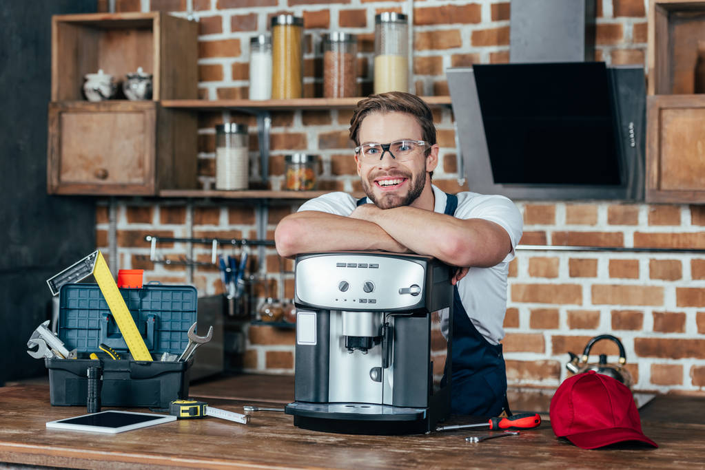 όμορφος νεαρός επισκευαστής ακουμπά στο μηχανή καφέ και να χαμογελά στη φωτογραφική μηχανή - Φωτογραφία, εικόνα