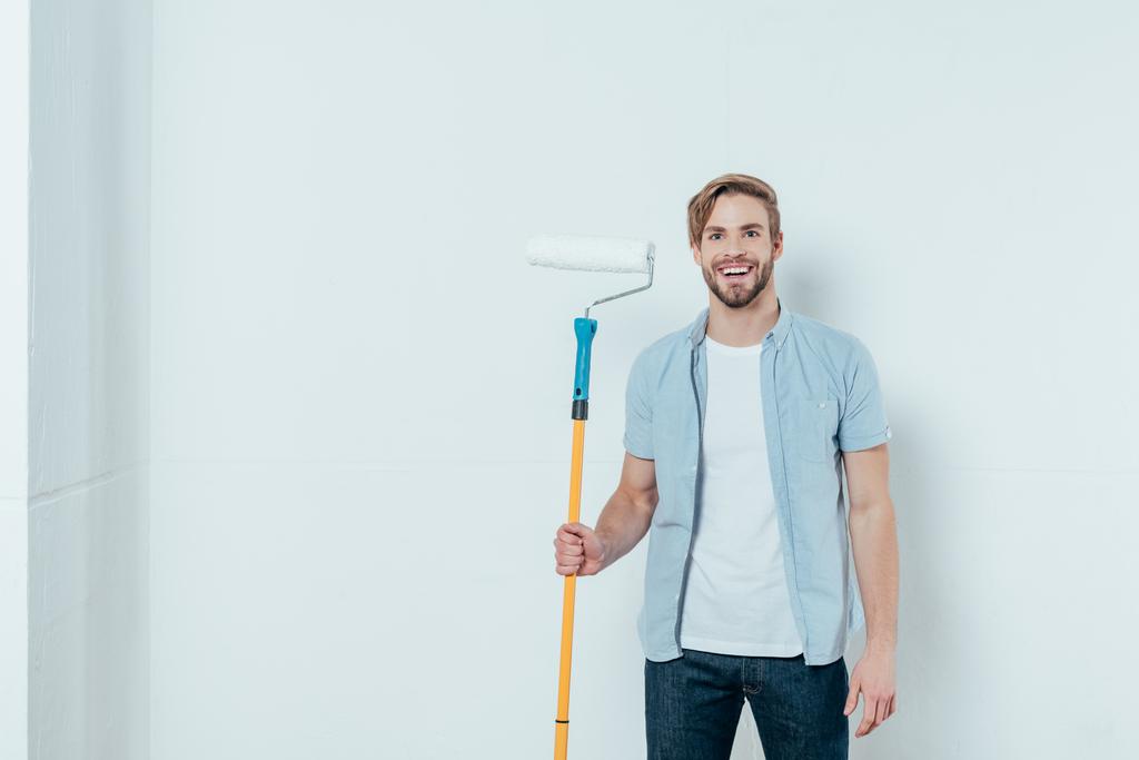 handsome young man holding paint roller and smiling at camera on grey - Photo, Image