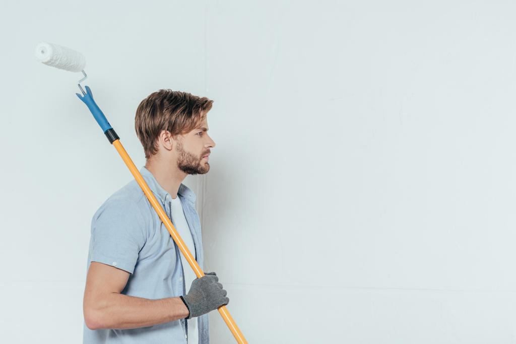 side view of young man holding paint roller and looking away on grey - Photo, Image
