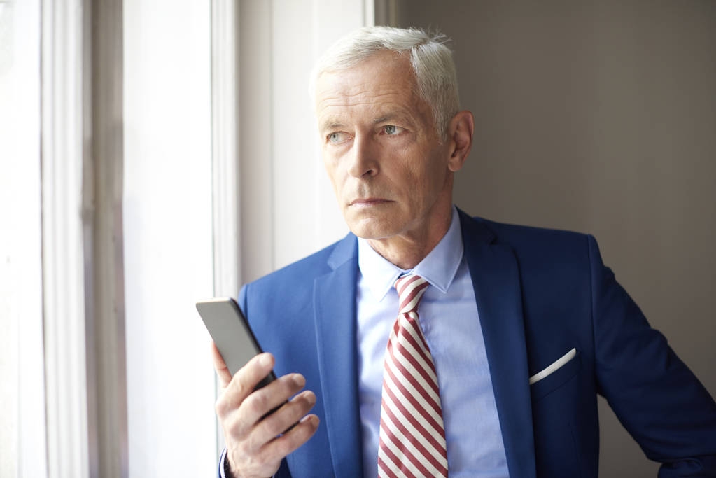 Hombre de negocios mayor con su teléfono móvil de pie en la ventana y mirando pensativamente
.  - Foto, Imagen