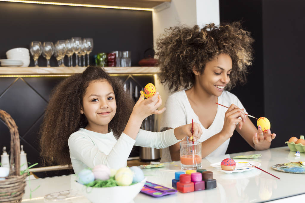 Prachtige Afrikaanse Amerikaanse vrouw en haar dochter kleuren paaseieren in de keuken - Foto, afbeelding