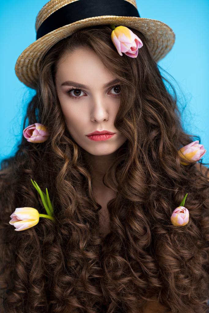 Kameraya bakarak uzun kıvırcık saçlarını çiçeklerle canotier şapkalı ciddi kadın - Fotoğraf, Görsel