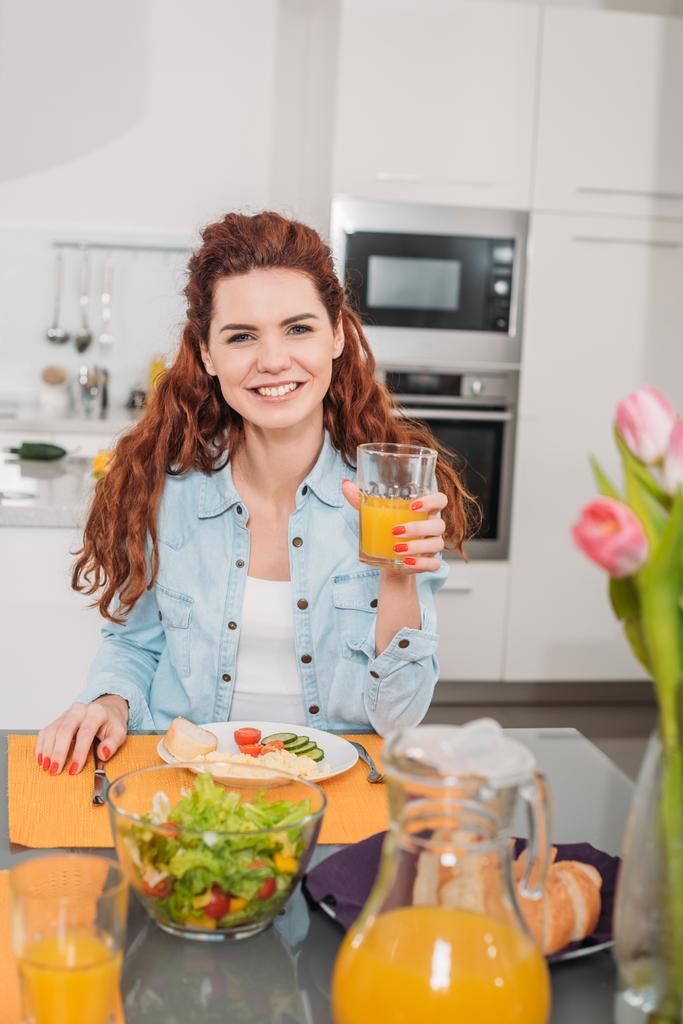 Uśmiechnięta dziewczyna siedzi przy stole z posiłkiem i trzymając szklankę soku pomarańczowego - Zdjęcie, obraz