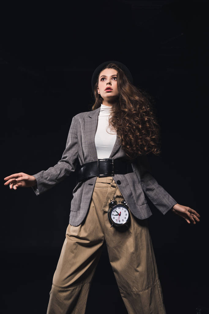 low angle view of fashionable shocked young woman with clock looking away isolated on black  - Photo, Image