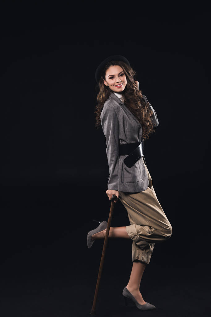 fashionable brunette woman posing with walking stick and smiling at camera isolated on black    - Photo, Image