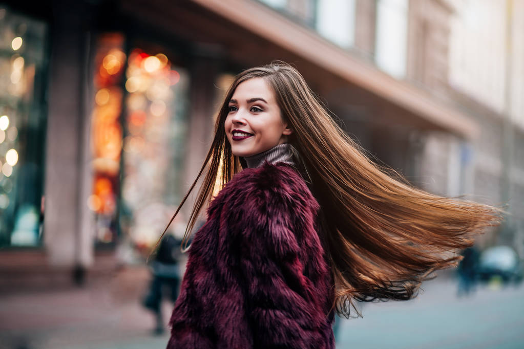 Jong mooi model meisje glimlacht en kijkt terug in de stad. Dynamisch jong meisje loopt door de straat.Haar fladdert in de wind. - Foto, afbeelding