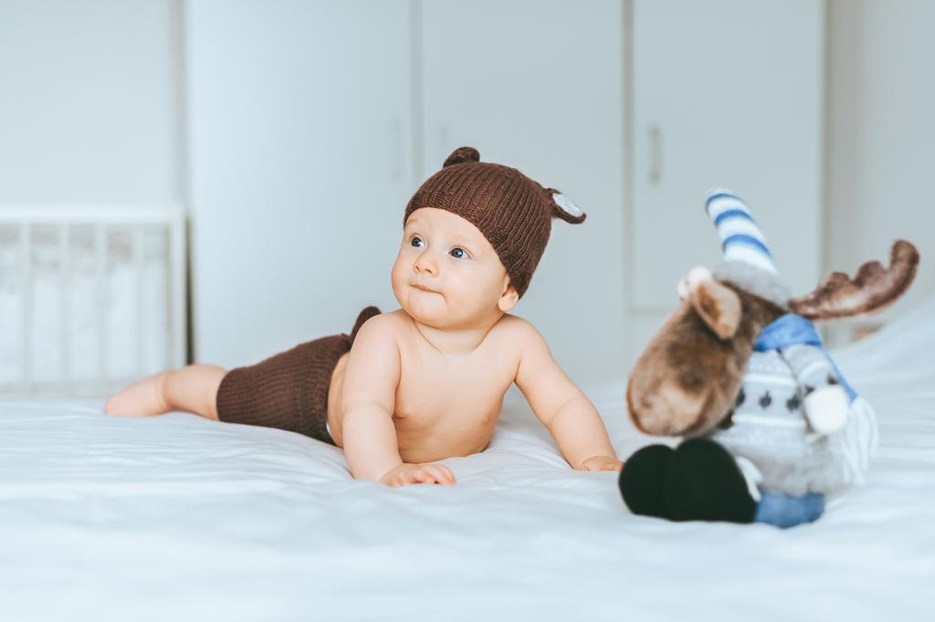 adorable infant child in knitted deer shorts and hat in bed with toy moose - Photo, Image