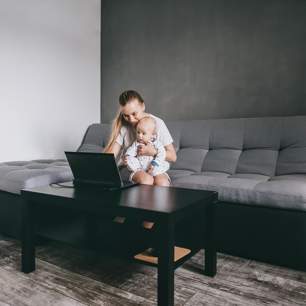 若い母親は自分の赤ちゃんとソファに座ってノート パソコンを使用して - 写真・画像