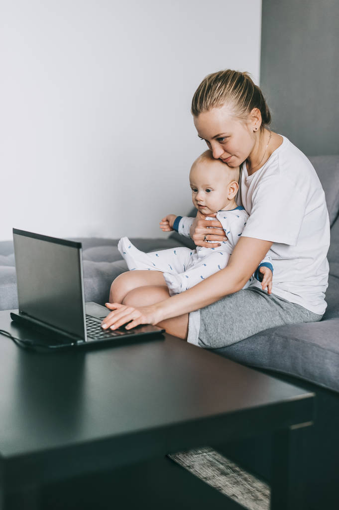 jovem mulher usando laptop enquanto carrega pouco bebê em casa
 - Foto, Imagem