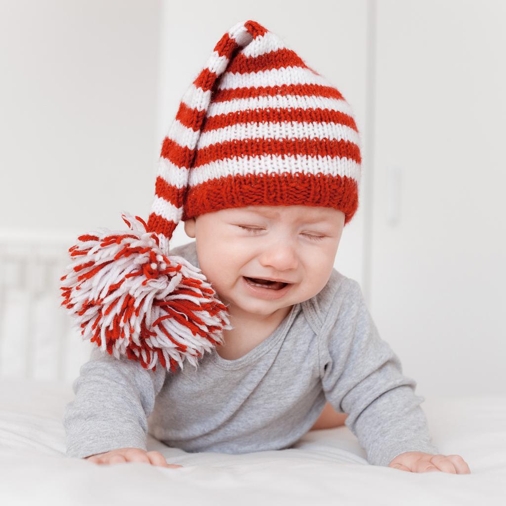 Porträt eines kleinen weinenden Säuglings mit lustiger Strickmütze auf dem Bett liegend  - Foto, Bild