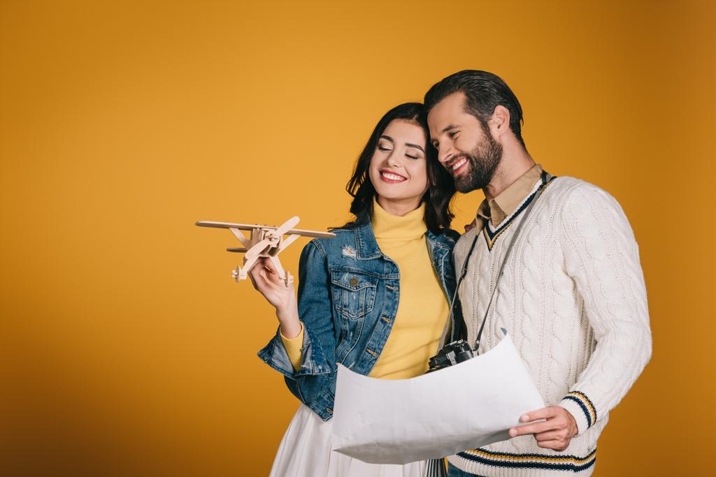 smiling couple looking at wooden plane isolated on yellow - Photo, Image