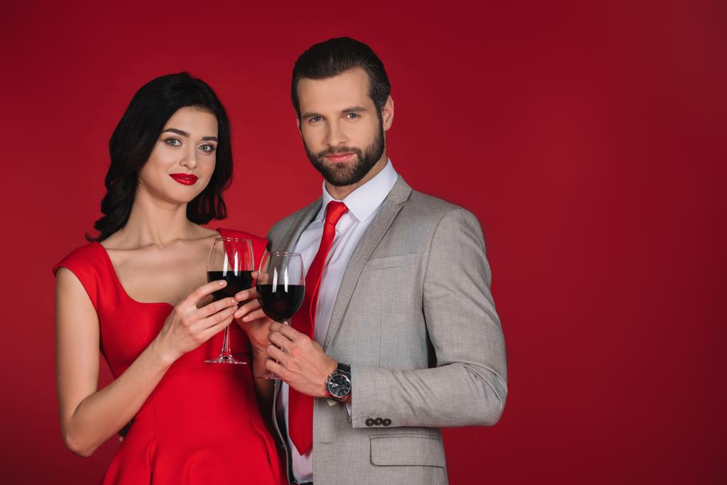couple holding glasses of wine and looking at camera isolated on red - Photo, Image