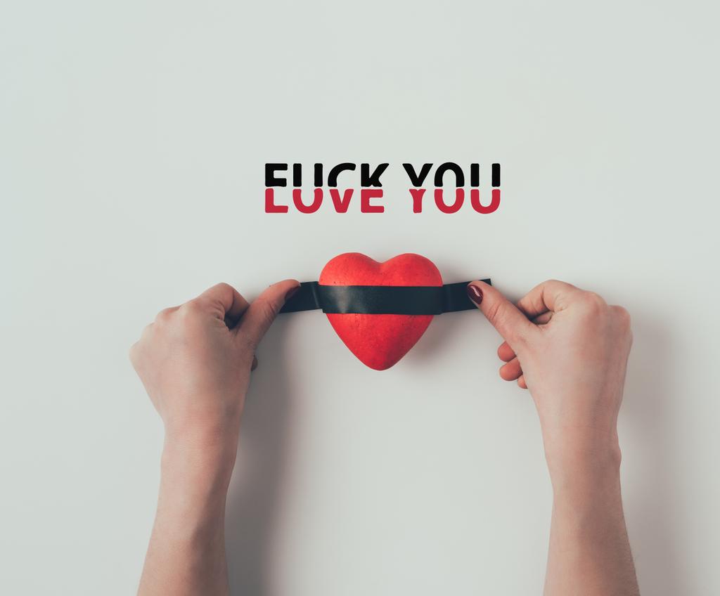 Cropped image of woman putting insulating tape on red heart with words love you fuck you on white - Foto, afbeelding