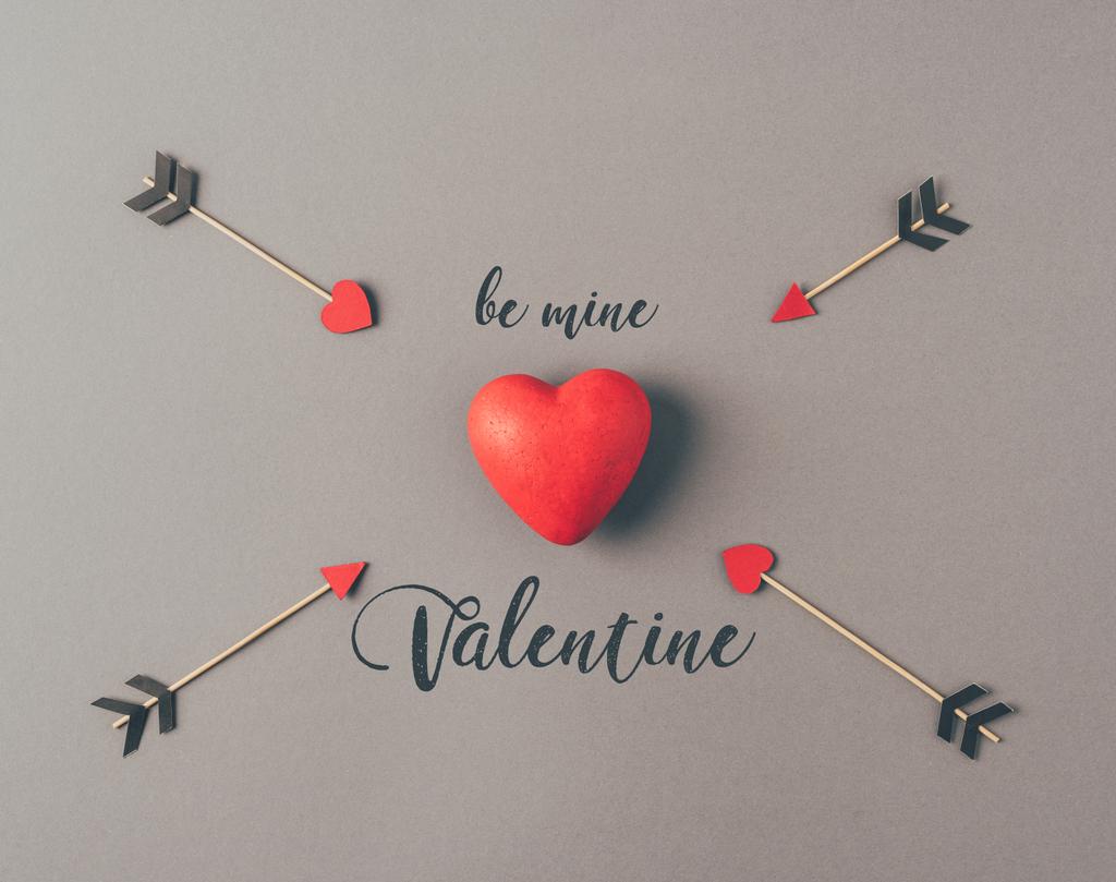 Bovenaanzicht van rood hart tussen vier pijlen en tekst worden mijne Valentine op grijs - Foto, afbeelding