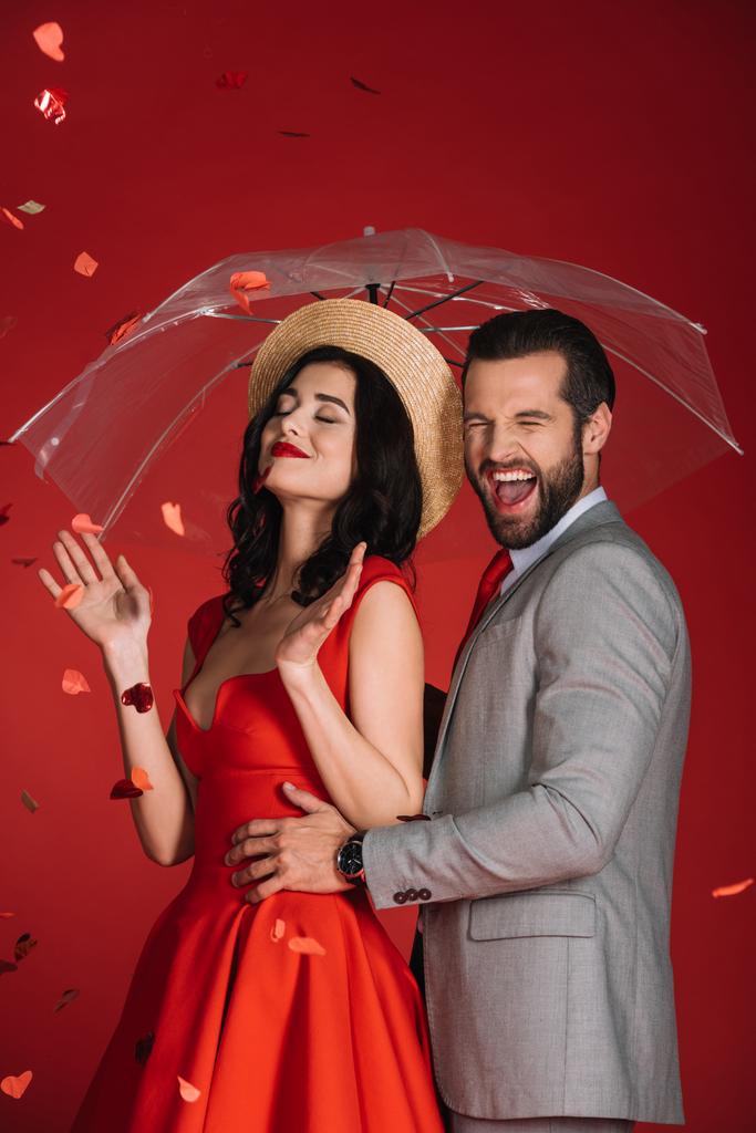 γελώντας ζευγάρι με ομπρέλα κάτω από πτώση κομφετί που απομονώνονται σε κόκκινο - Φωτογραφία, εικόνα
