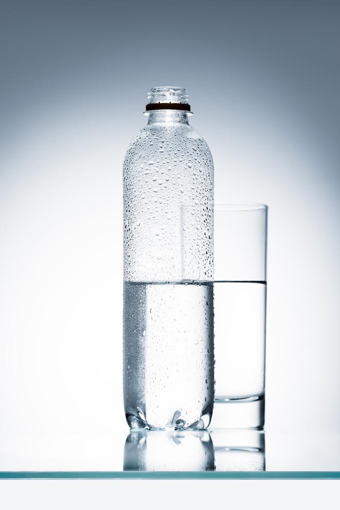 plastic fles en glas water op reflecterend oppervlak - Foto, afbeelding