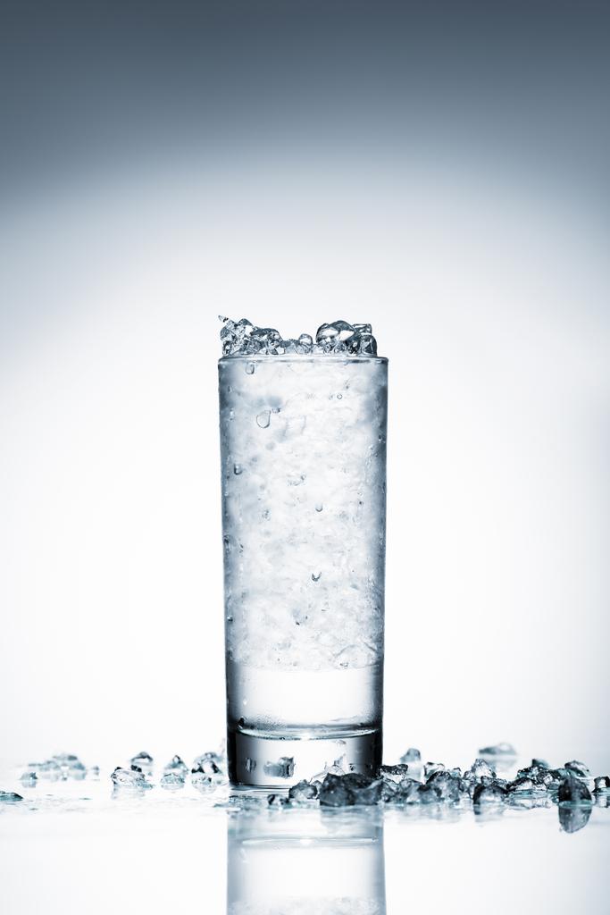 ποτήρι κρύο νερό με θρυμματισμένο πάγο σε ανακλαστική επιφάνεια σε λευκό  - Φωτογραφία, εικόνα
