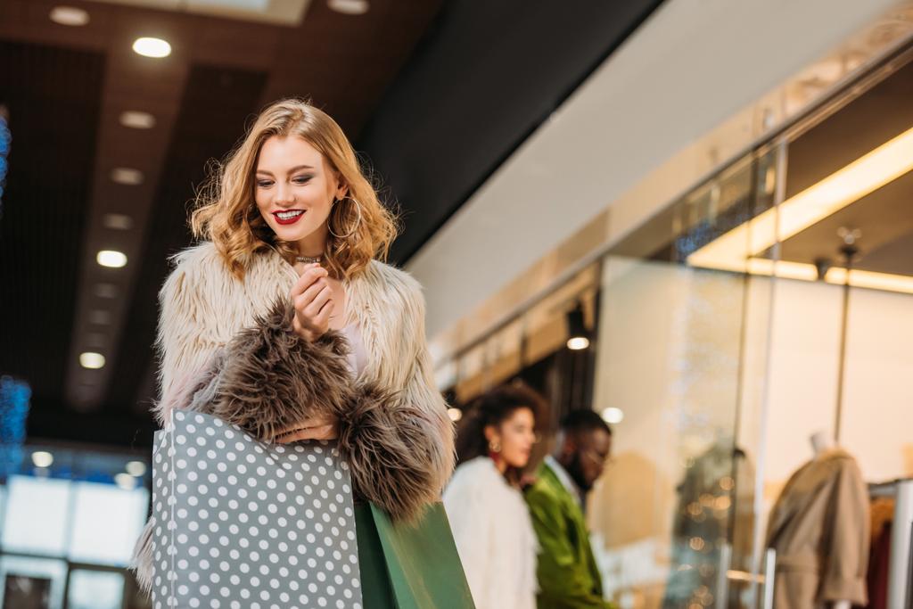 sorrindo elegante jovem mulher em casaco de pele segurando sacos de compras enquanto caminhava no shopping
 - Foto, Imagem