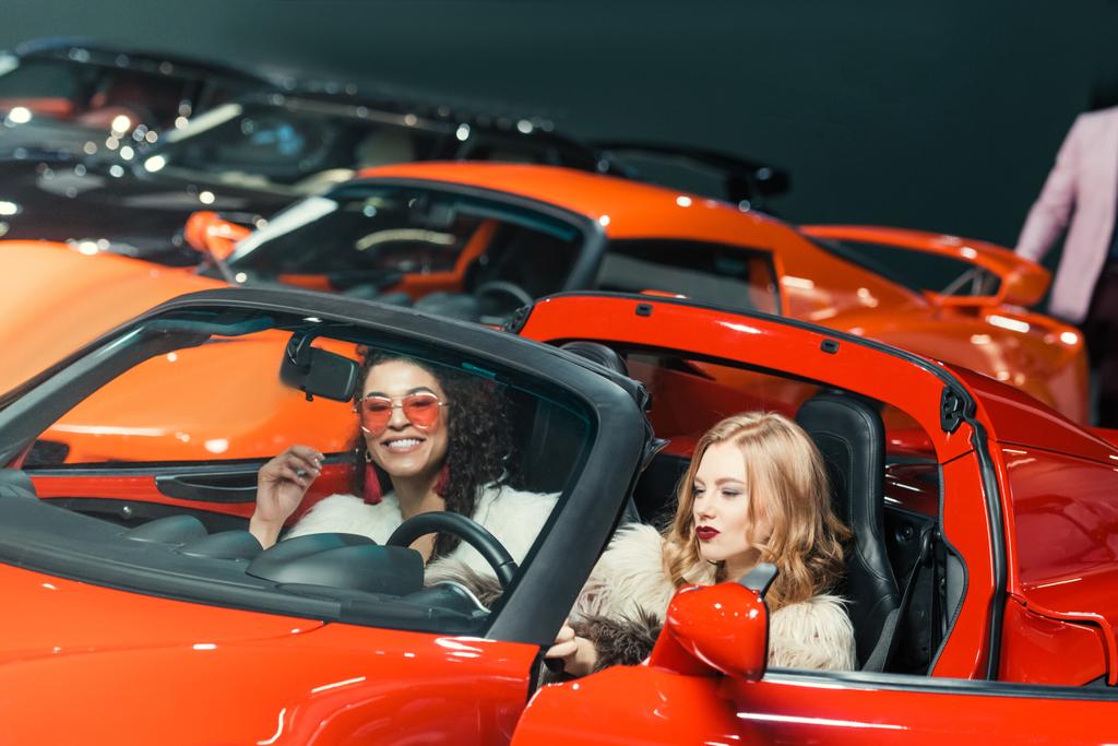 mujeres multiétnicas sonrientes de moda sentadas en un coche rojo de lujo
 - Foto, Imagen