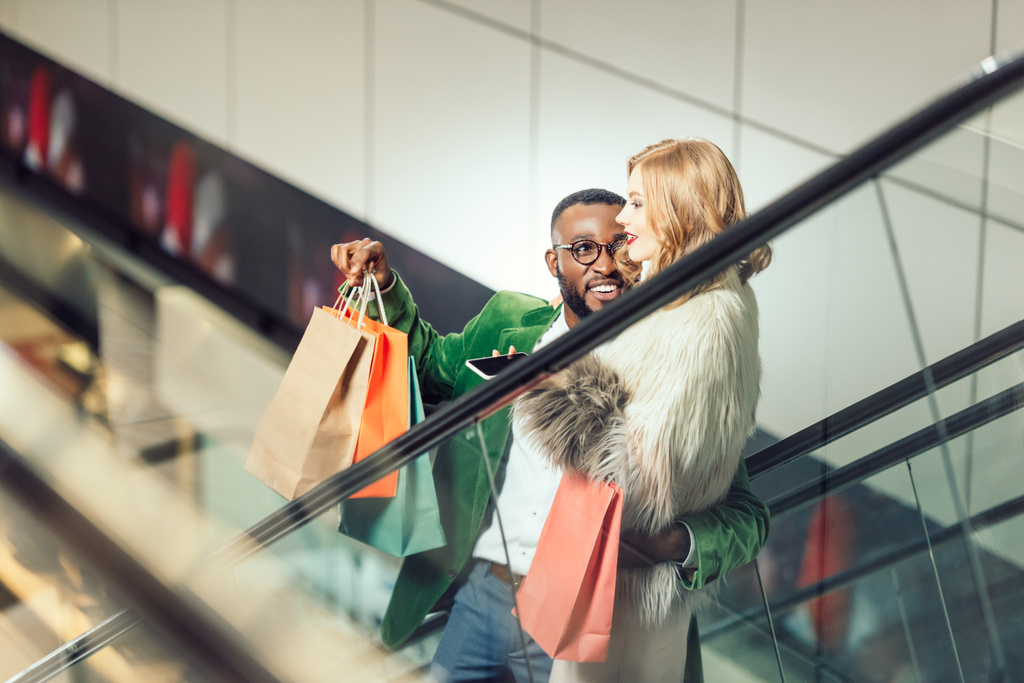 feliz joven interracial pareja hablando mientras montando escaleras mecánicas en el centro comercial
 - Foto, Imagen