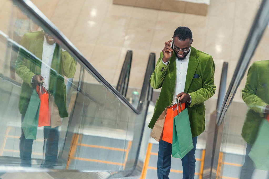 homme élégant souriant avec sac à provisions parlant par téléphone sur l'escalator au centre commercial
 - Photo, image