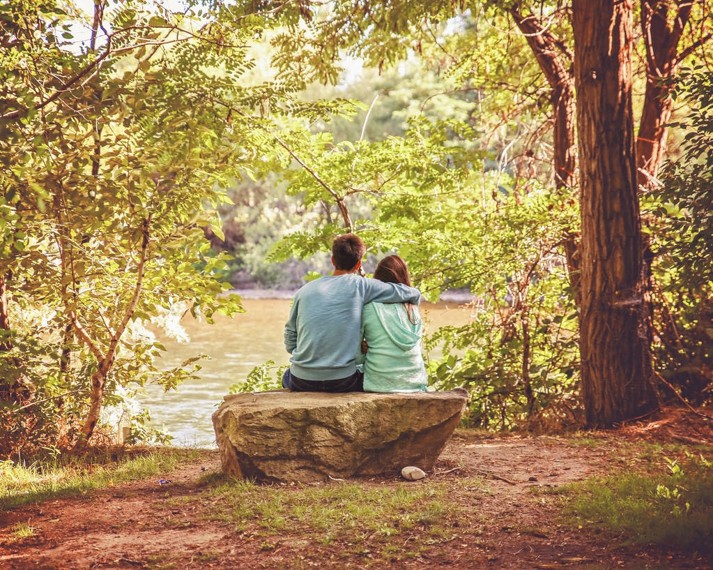 a couple in love sitting on a river bank with the man draping his arm over the woman on a warm summer day - Photo, Image