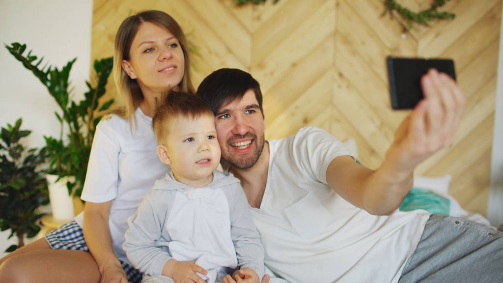 Smiling parents with baby taking selfie family photo on bed at home - Photo, Image
