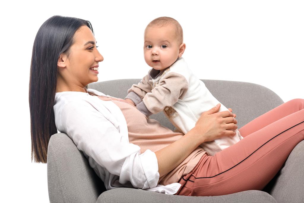 lachende jonge moeder rust op arm stoel samen met kleine baby geïsoleerd op wit - Foto, afbeelding