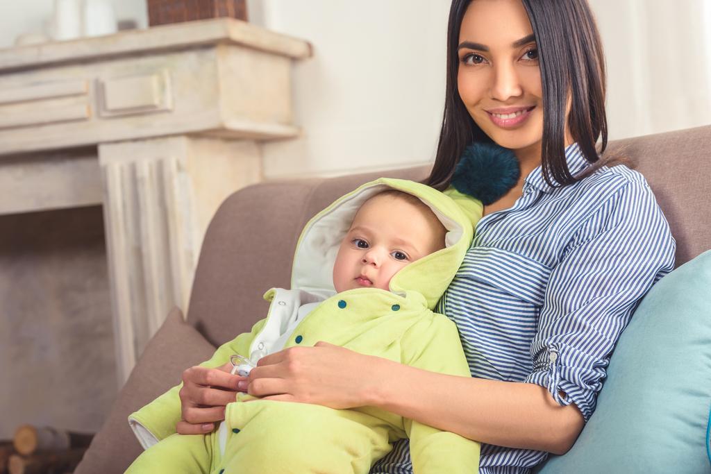 πορτρέτο του χαμογελώντας η μητέρα με το μωρό στα χέρια ακουμπά στον καναπέ στο σπίτι - Φωτογραφία, εικόνα