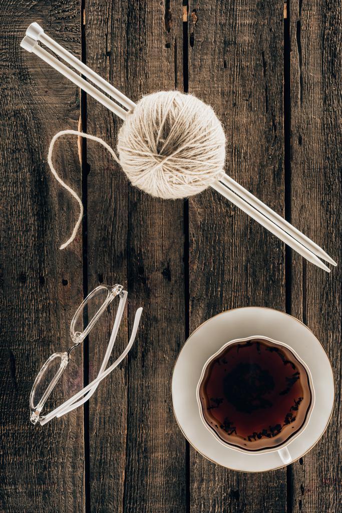 Draufsicht auf Stricknadeln, Garn, Tasse Tee und Brille auf Holzgrund - Foto, Bild