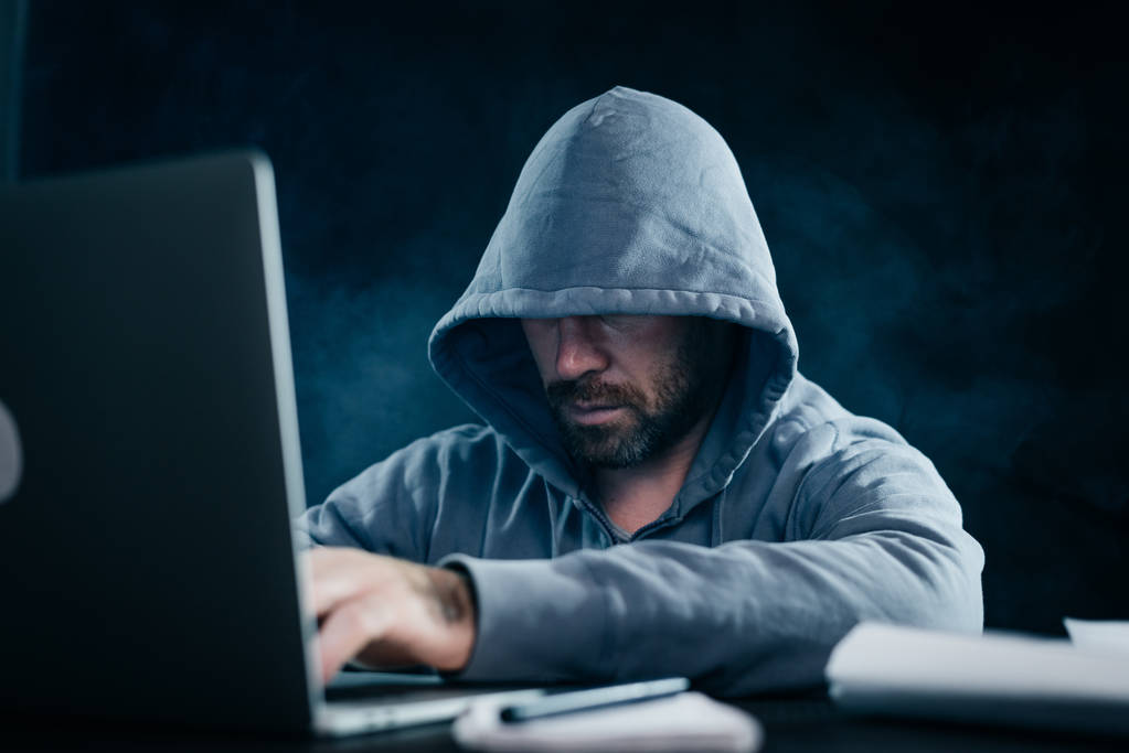 хлопець хакер з бородою в светрі з капюшоном сидить за ноутбуком
 - Фото, зображення