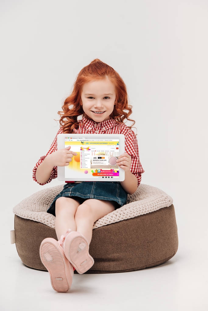 Suloinen pieni tyttö hymyilee kameralle pitäen digitaalinen tabletti aliexpress verkkosivuilla ruudulla eristetty harmaa
 - Valokuva, kuva