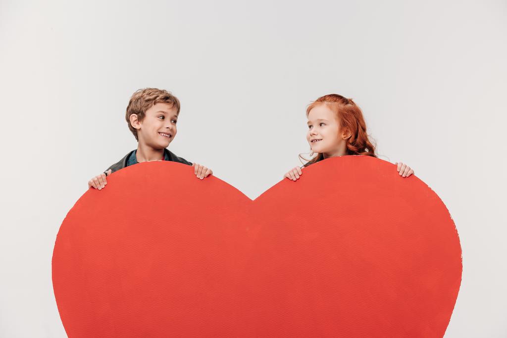 χαμογελώντας λίγο παιδιά ζευγάρι πίσω από την μεγάλη κόκκινη καρδιά που απομονώνονται σε γκρι - Φωτογραφία, εικόνα