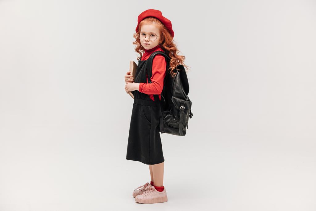 puolella näkymä kaunis pieni koulutyttö punainen baskeri kirja ja reppu eristetty harmaa
 - Valokuva, kuva