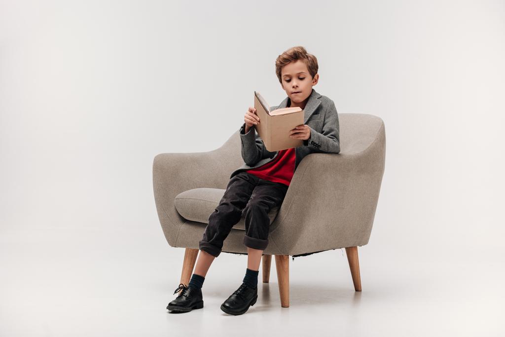 focused little schoolboy reading book in armchair - Photo, Image