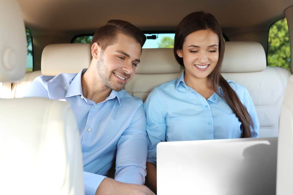 Hermosa pareja joven sentada en el asiento trasero con el ordenador portátil en el coche
 - Foto, Imagen