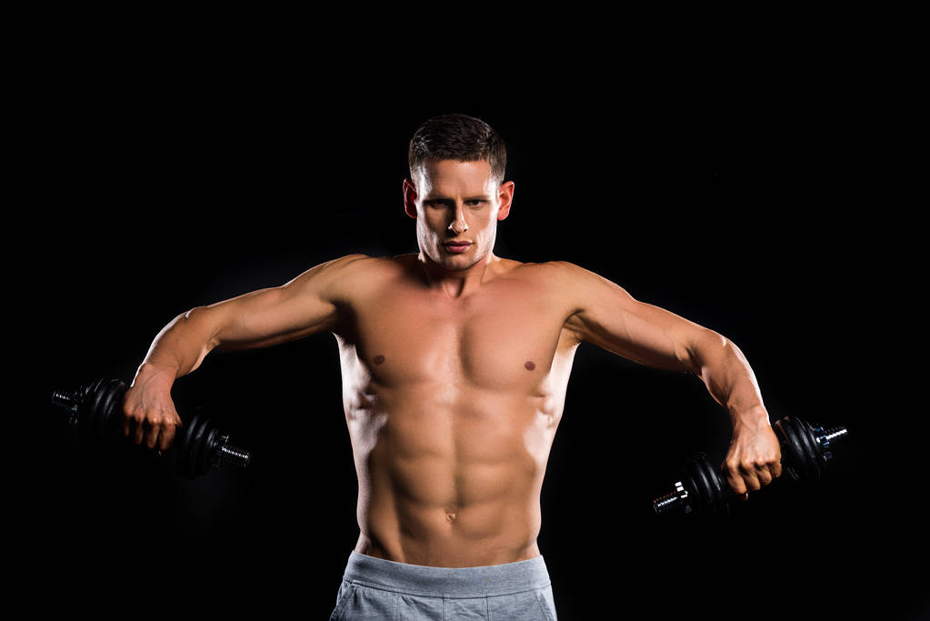 young muscular shirtless sportsman holding dumbbells and looking at camera isolated on black  - Photo, Image