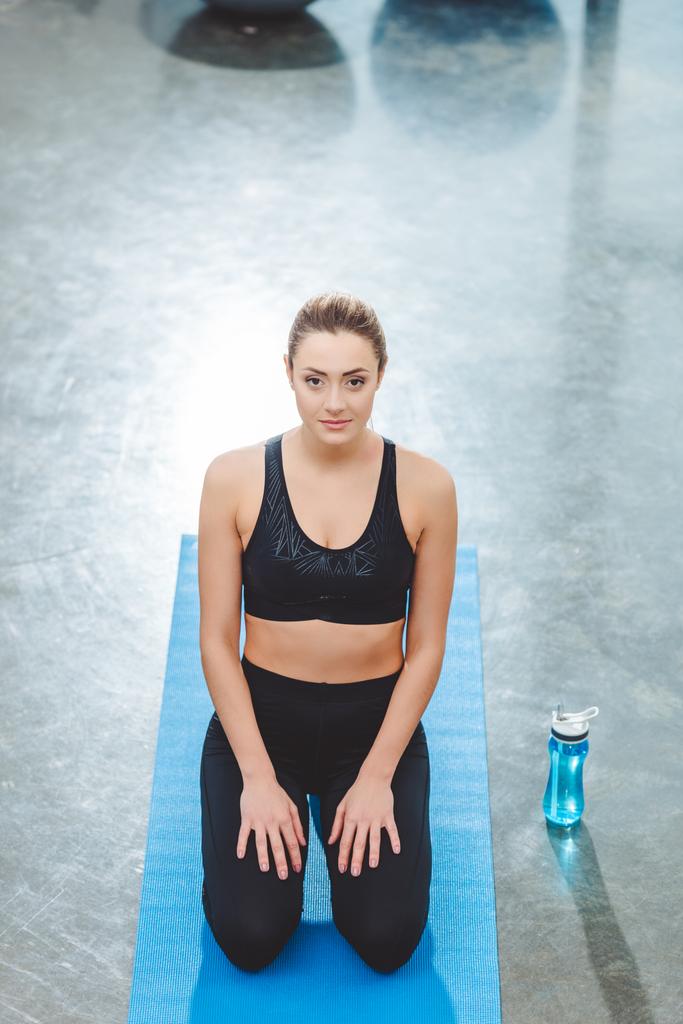 hoge hoekmening van jonge Sportvrouw zittend op yoga mat en kijken naar de camera in de fitness-studio - Foto, afbeelding