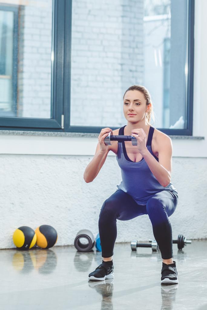 bodur egzersiz yaparak ve spor salonunda dumbbell tutarak atletik genç kadın - Fotoğraf, Görsel