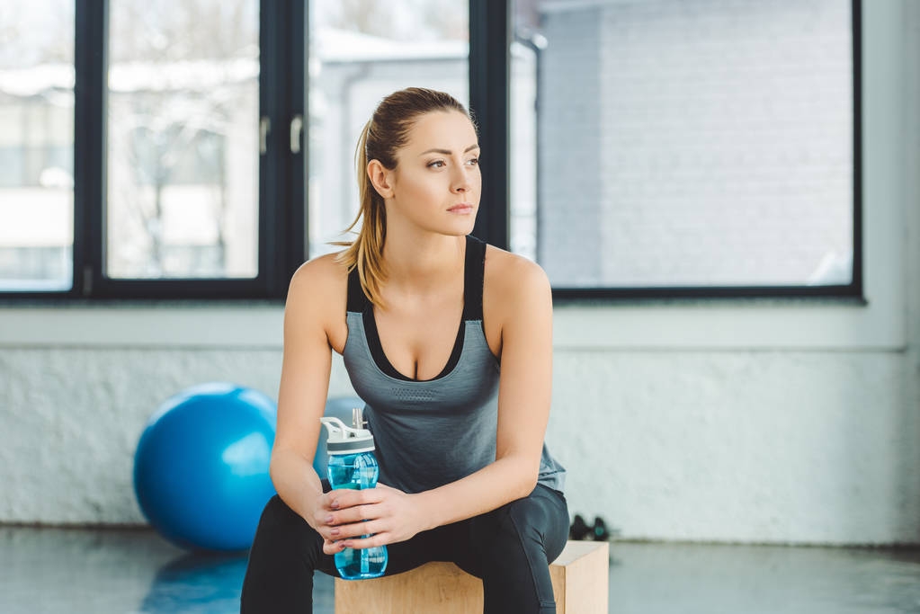 πορτρέτο του συλλογισμένος αθλήτρια με μπουκάλι νερό κοιτάζοντας μακριά στο γυμναστήριο - Φωτογραφία, εικόνα