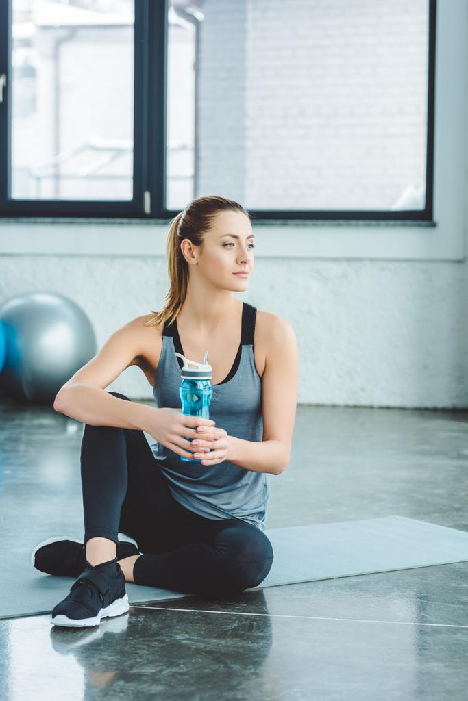 спортсменка с бутылкой воды отдыхает на коврике после тренировки в тренажерном зале
 - Фото, изображение