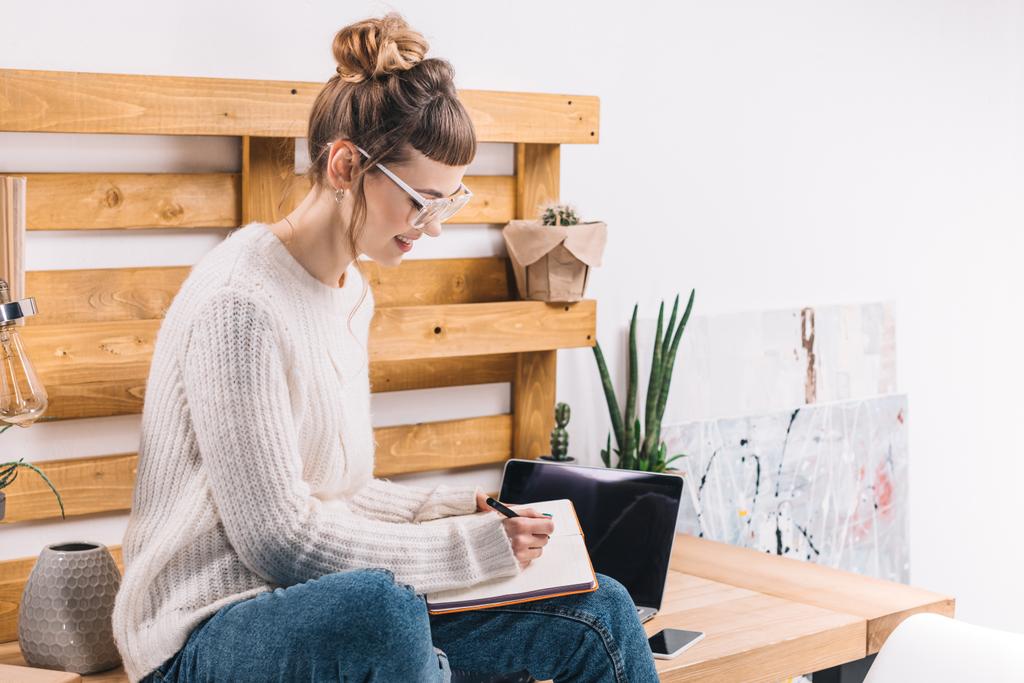 smiling girl sitting on table in office and writing something to notebook  - Photo, Image