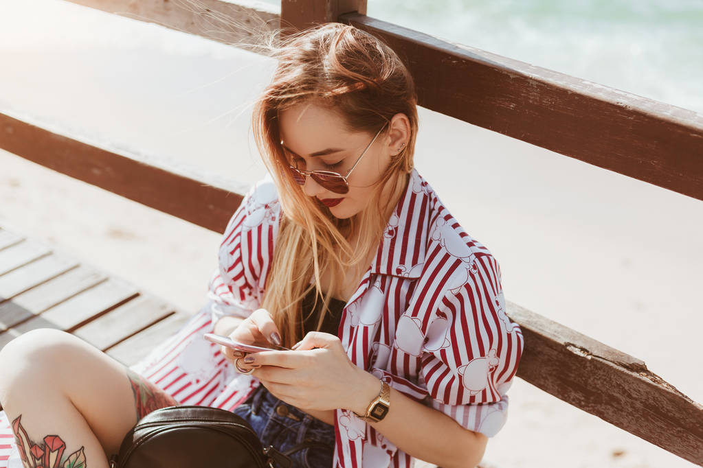 Blick aus der Vogelperspektive auf junge Frau, die am Strand auf einem Steg sitzt und ihr Smartphone benutzt - Foto, Bild