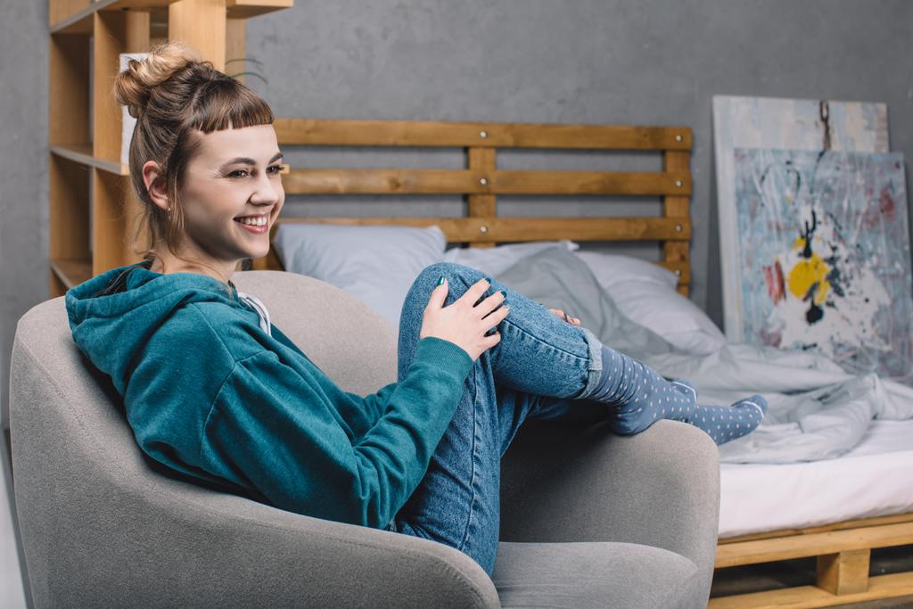 улыбающаяся девушка, сидящая на кресле в спальне и отводящая взгляд
 - Фото, изображение